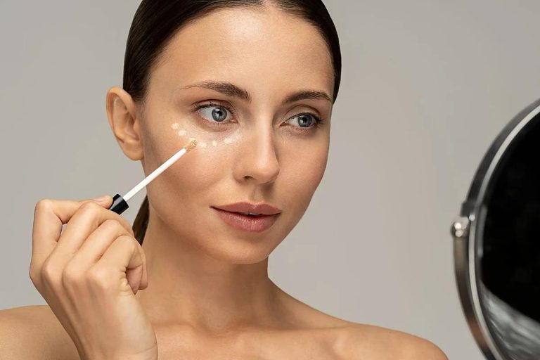 Mujer aplicando corrector de ojeras en pequeños puntos bajo el ojo mientras se mira en un espejo.