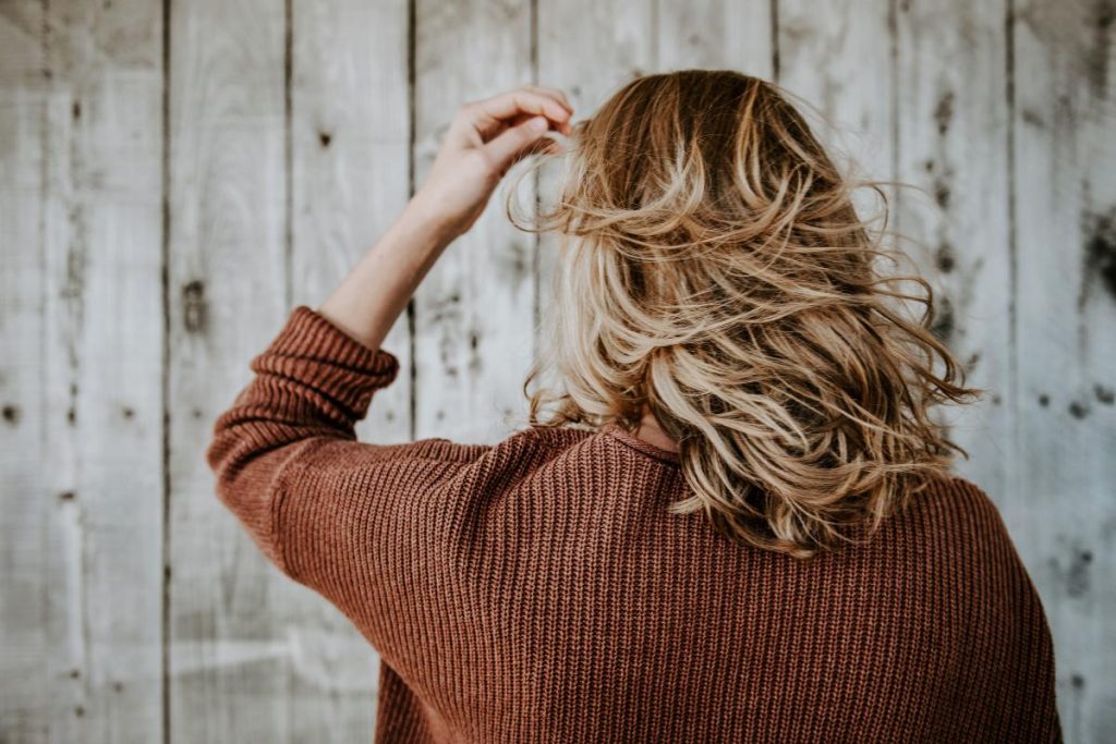 Mujer de espaldas con melena ondulada castaña y mechas rubias frente a una pared de madera.